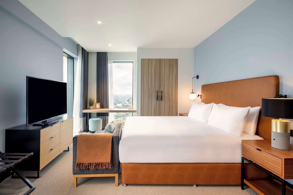 Una habitación de hotel moderna con una cama grande, televisión, escritorio y ventana con vista a la ciudad.