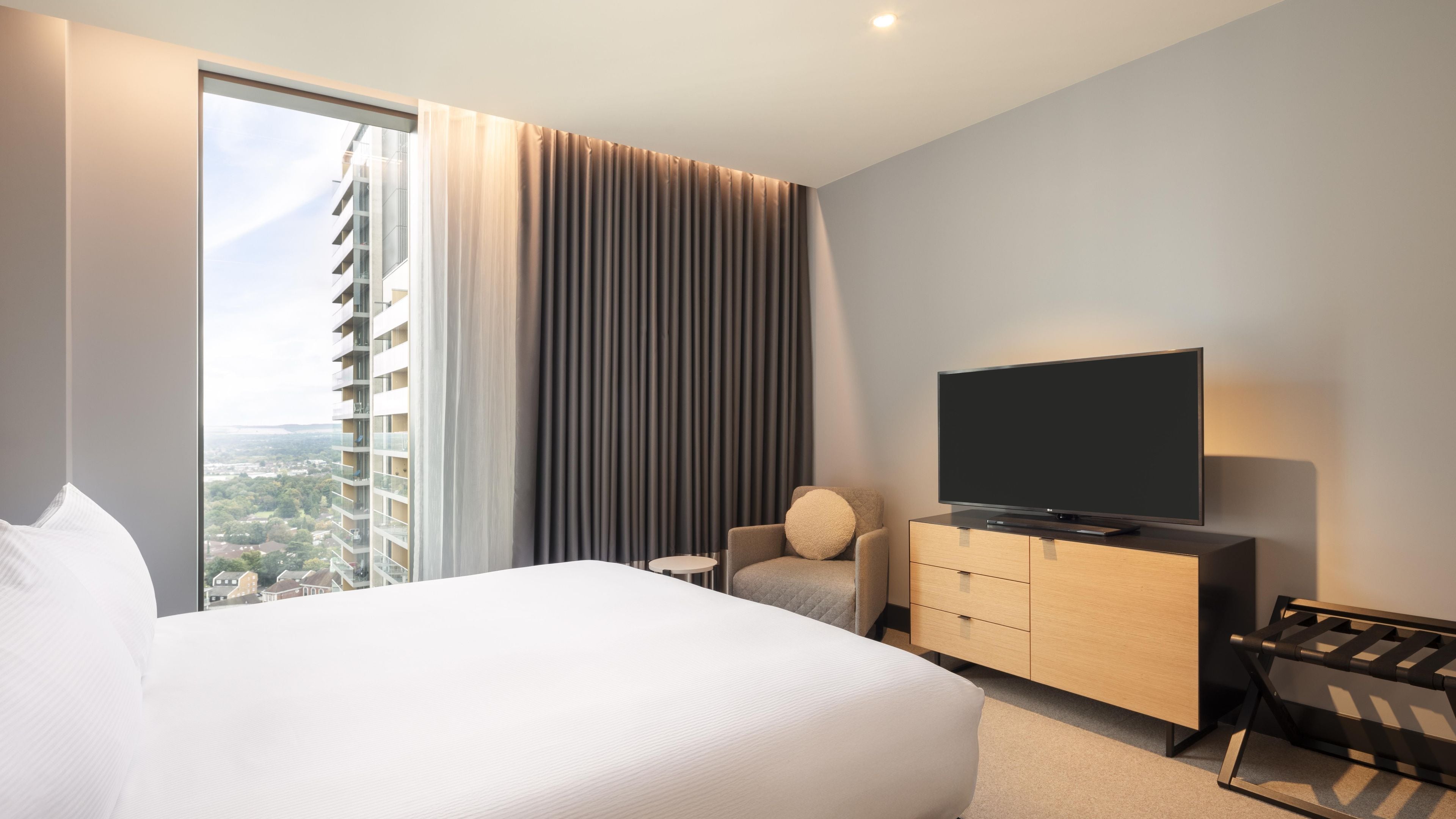 Habitación de hotel moderna con una cama grande, un televisor de pantalla plana, un aparador de madera y vistas a un paisaje urbano por la ventana.