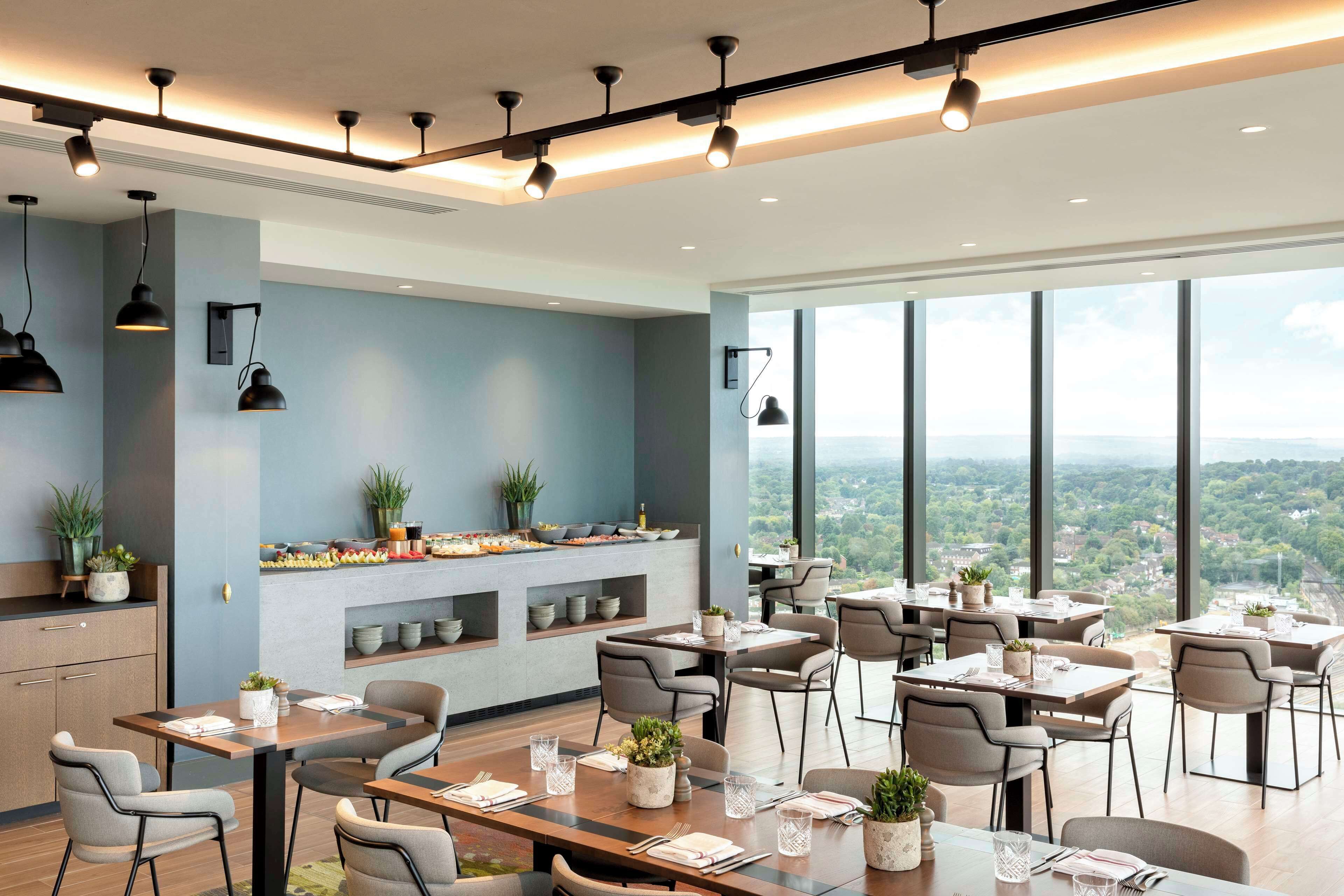 A modern dining area with multiple tables and chairs, a kitchen counter with plants, and large windows offering a city view.