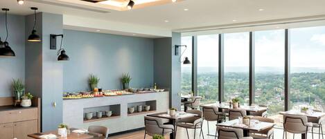 A modern dining area with multiple tables and chairs, a kitchen counter with plants, and large windows offering a city view.