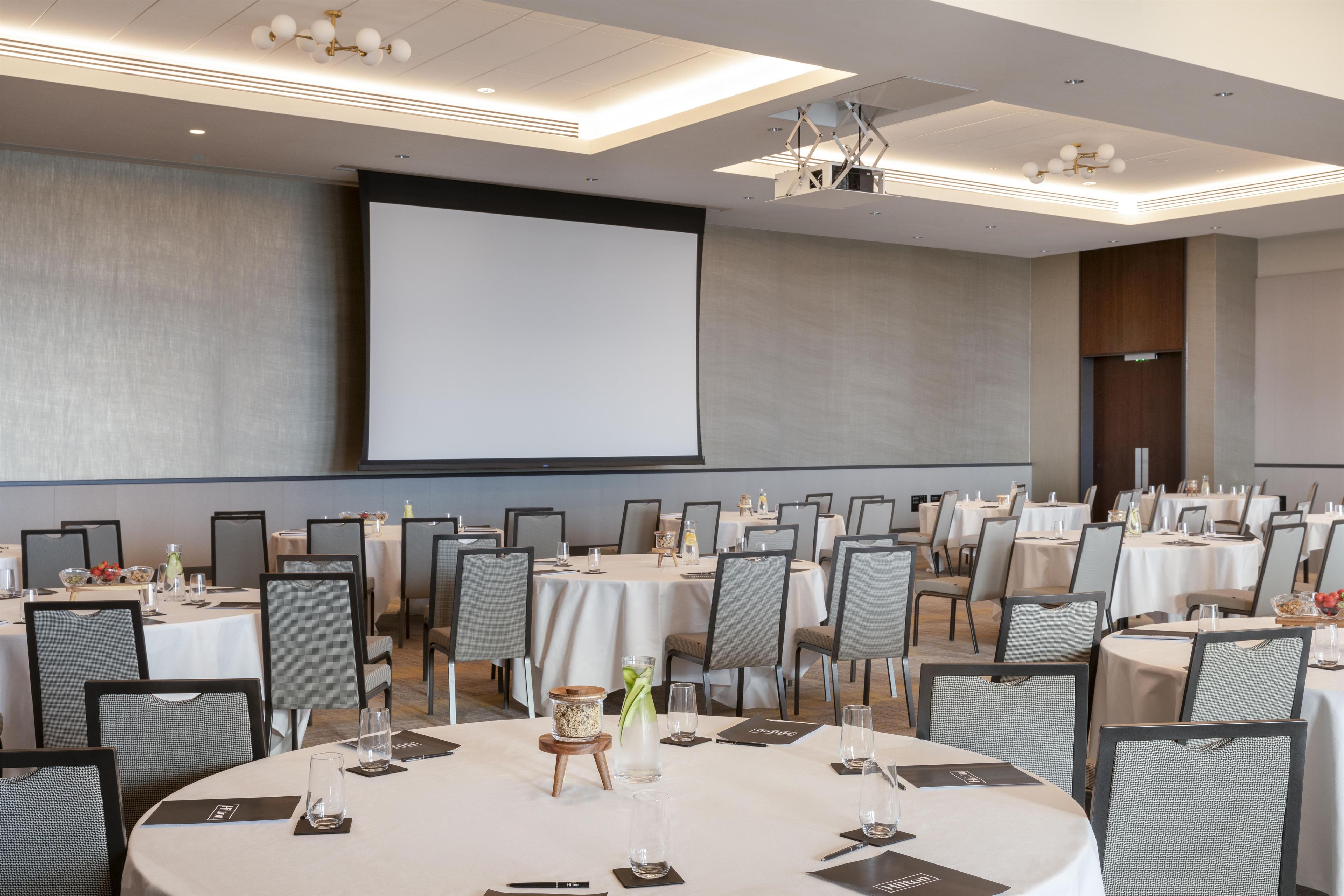 Un salón de conferencias con varias mesas, sillas y una pantalla grande para presentaciones.