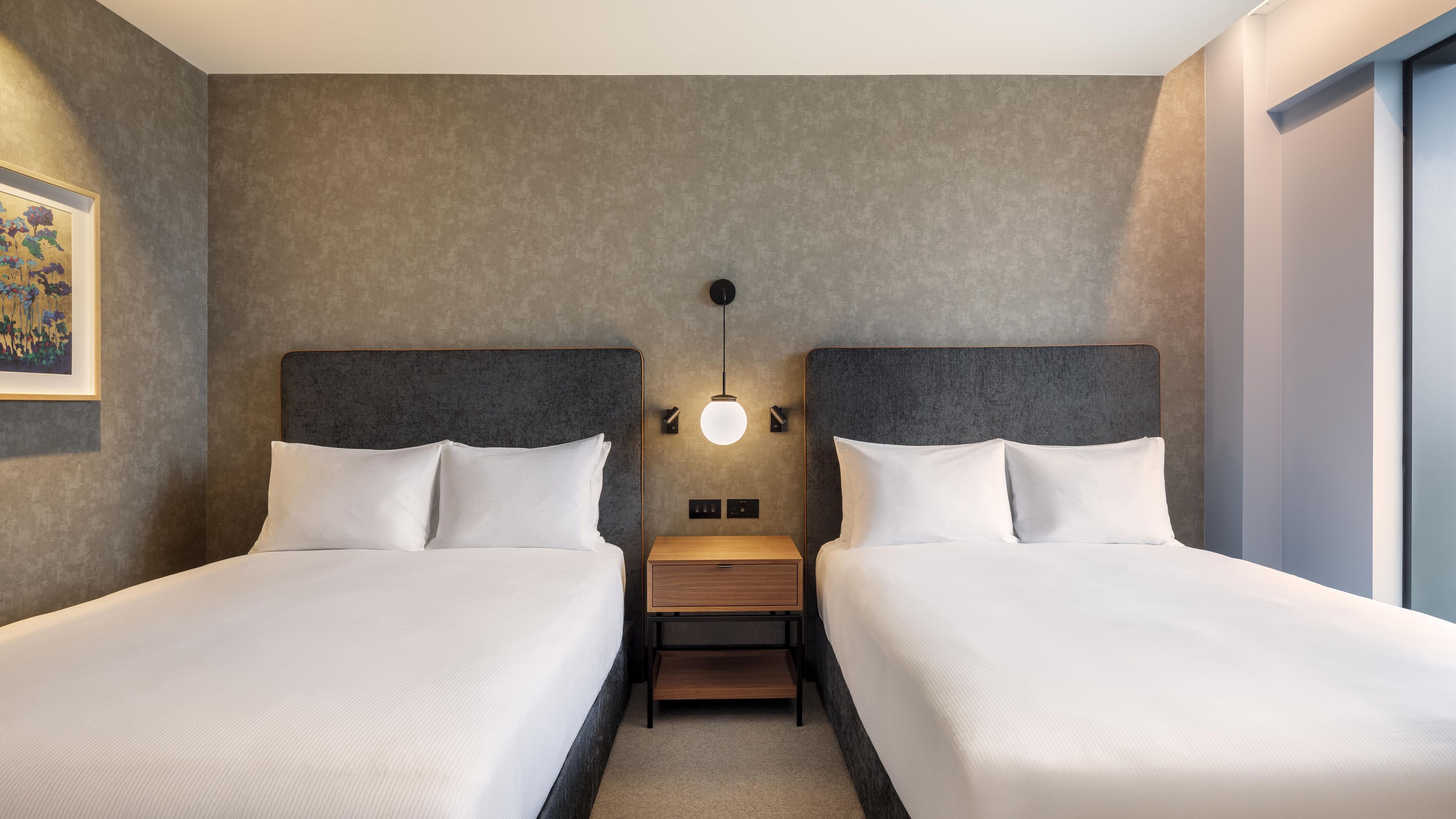 A hotel room with two beds, a nightstand, a wall-mounted lamp, and a framed picture.