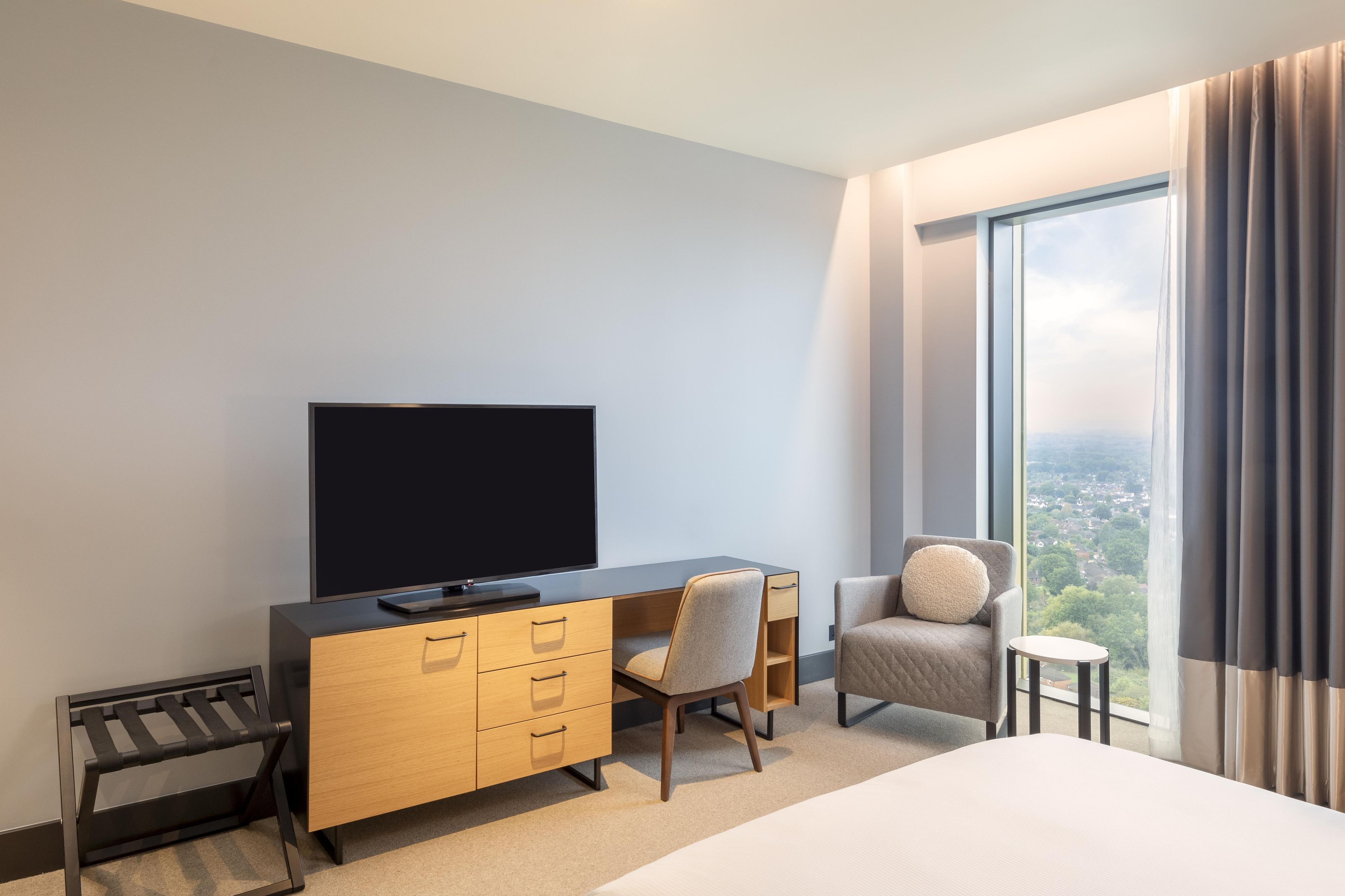 Habitación de hotel con cama, televisor en una cómoda de madera, una silla y vistas a un paisaje urbano a través de grandes ventanales.