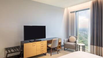 Habitación de hotel con cama, televisor en una cómoda de madera, una silla y vistas a un paisaje urbano a través de grandes ventanales.