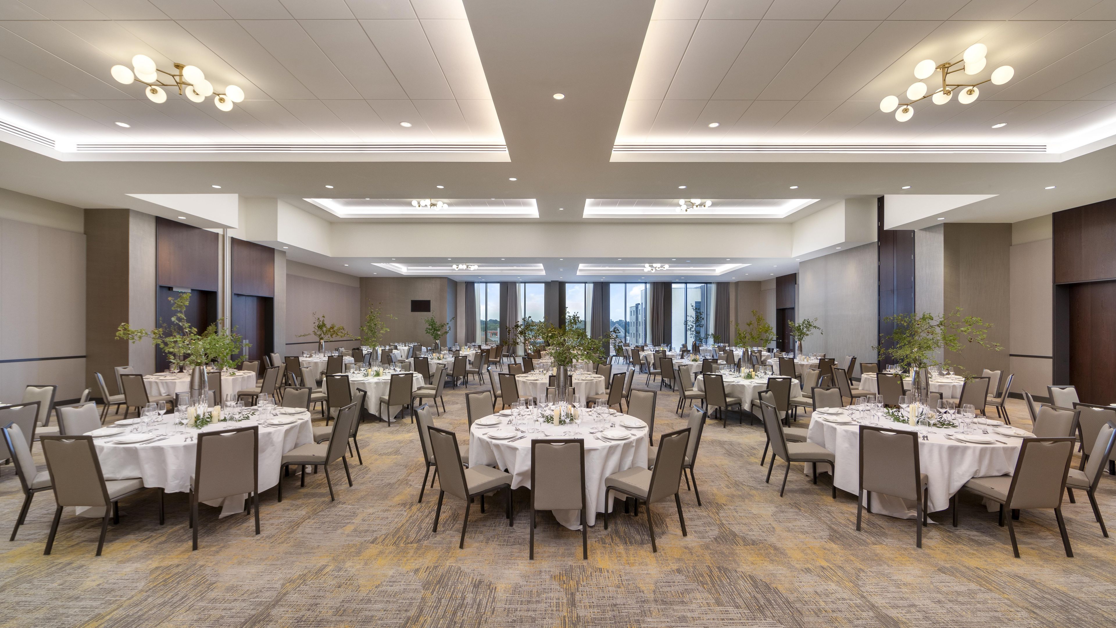 A large banquet hall with round tables set for dining, featuring a carpeted floor and a high ceiling with recessed lighting.