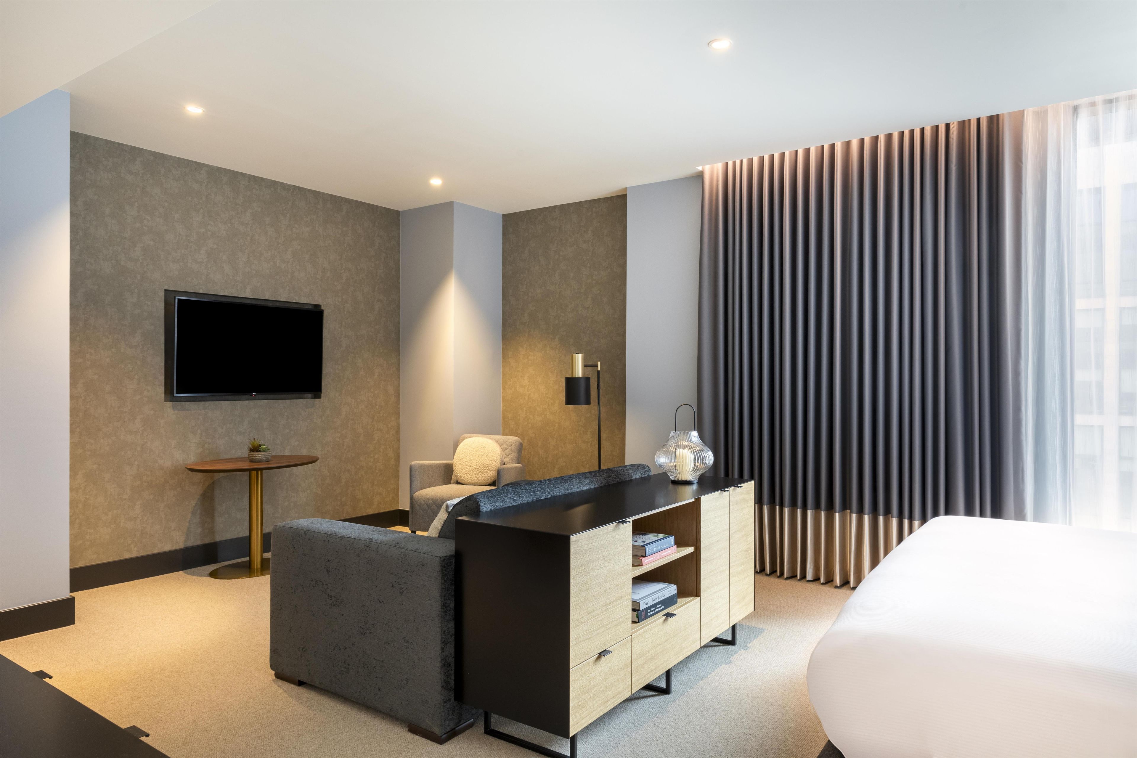 A modern hotel room with a flat-screen TV mounted on the wall, a sofa, a small side table with a lamp, and a large window with curtains.