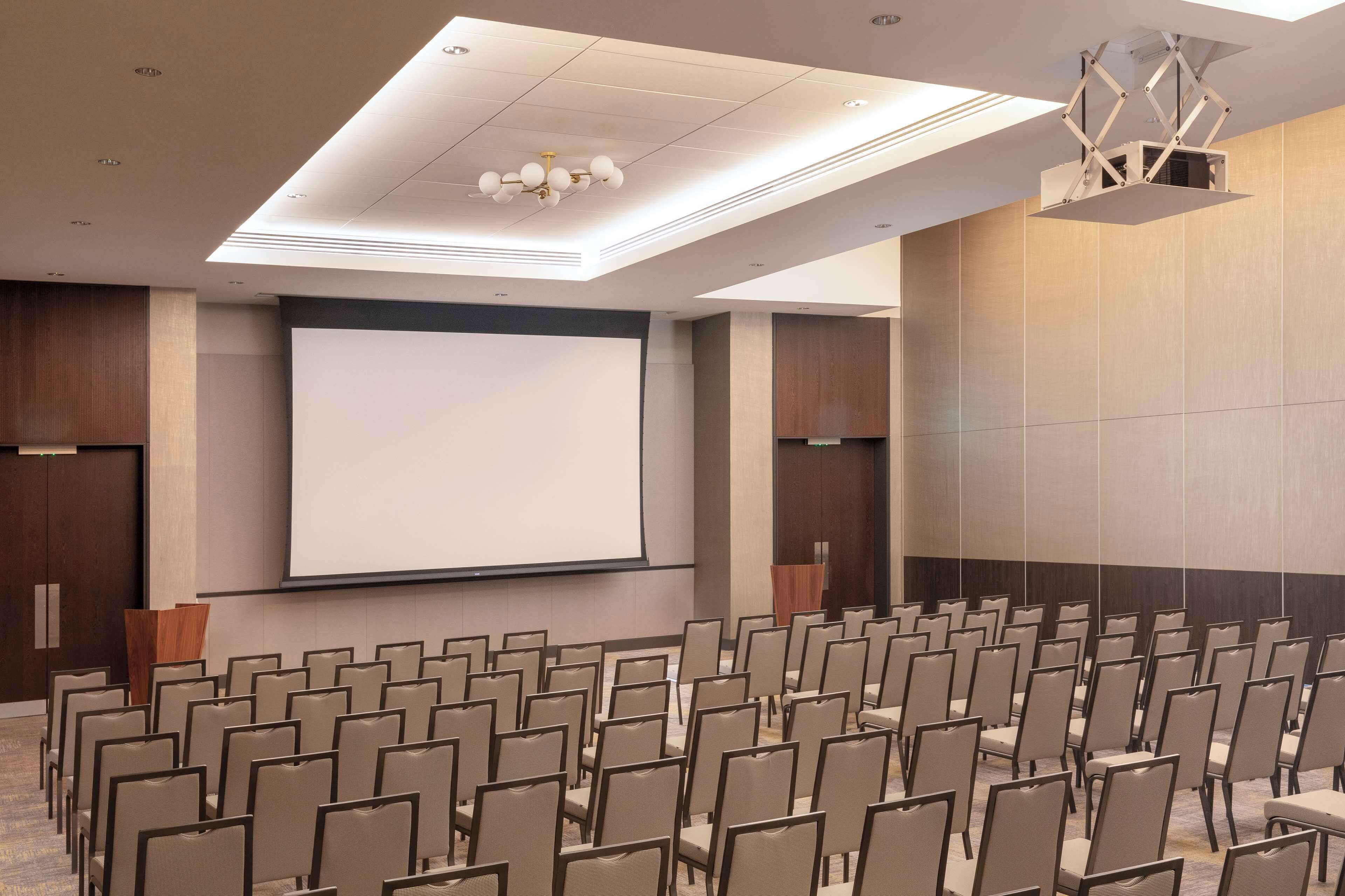 Un salón de conferencias con filas de sillas, una pantalla grande y una lámpara en el techo.