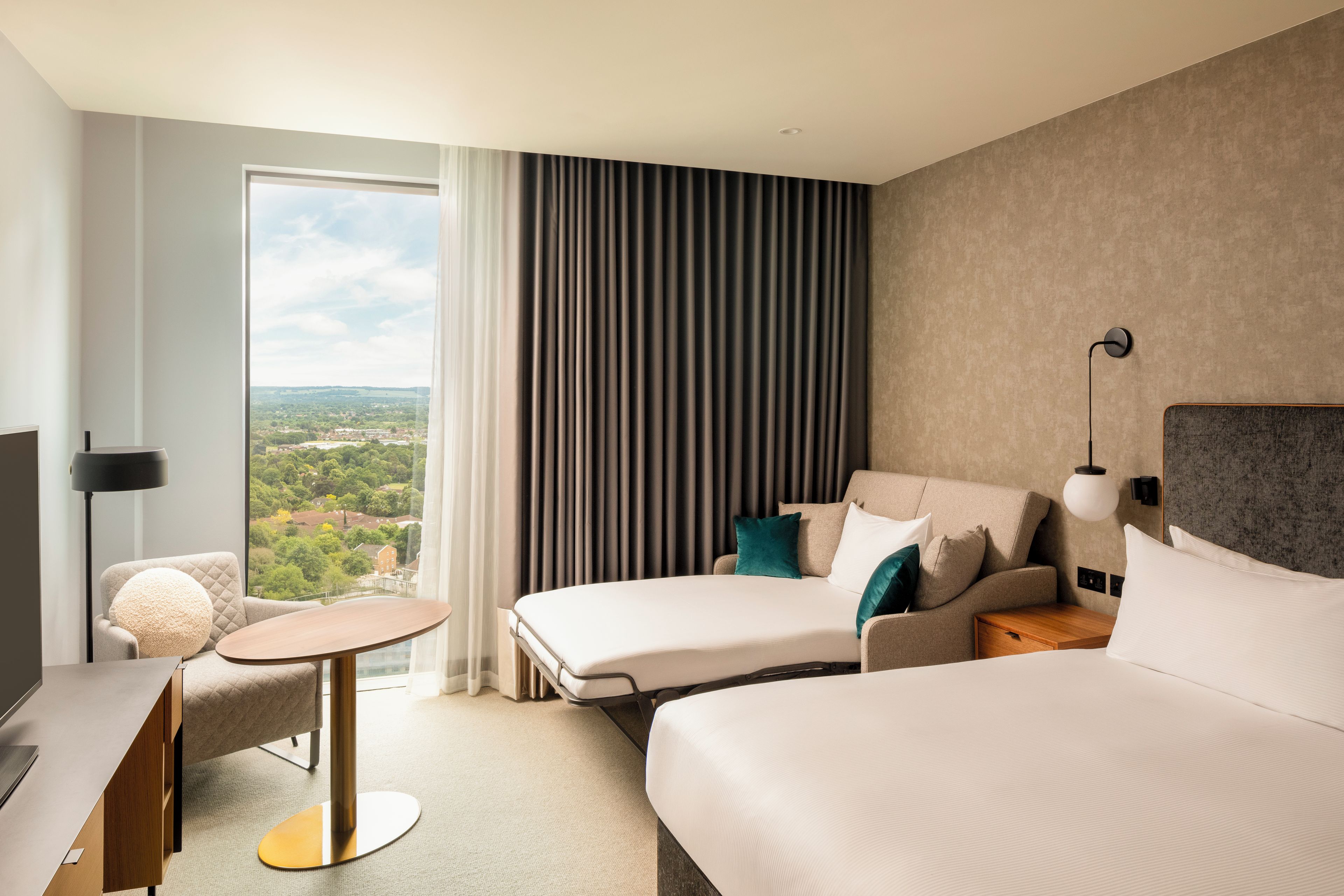 Quarto de hotel com cama, escrivaninha, cadeira, luminária e vista para a cidade.