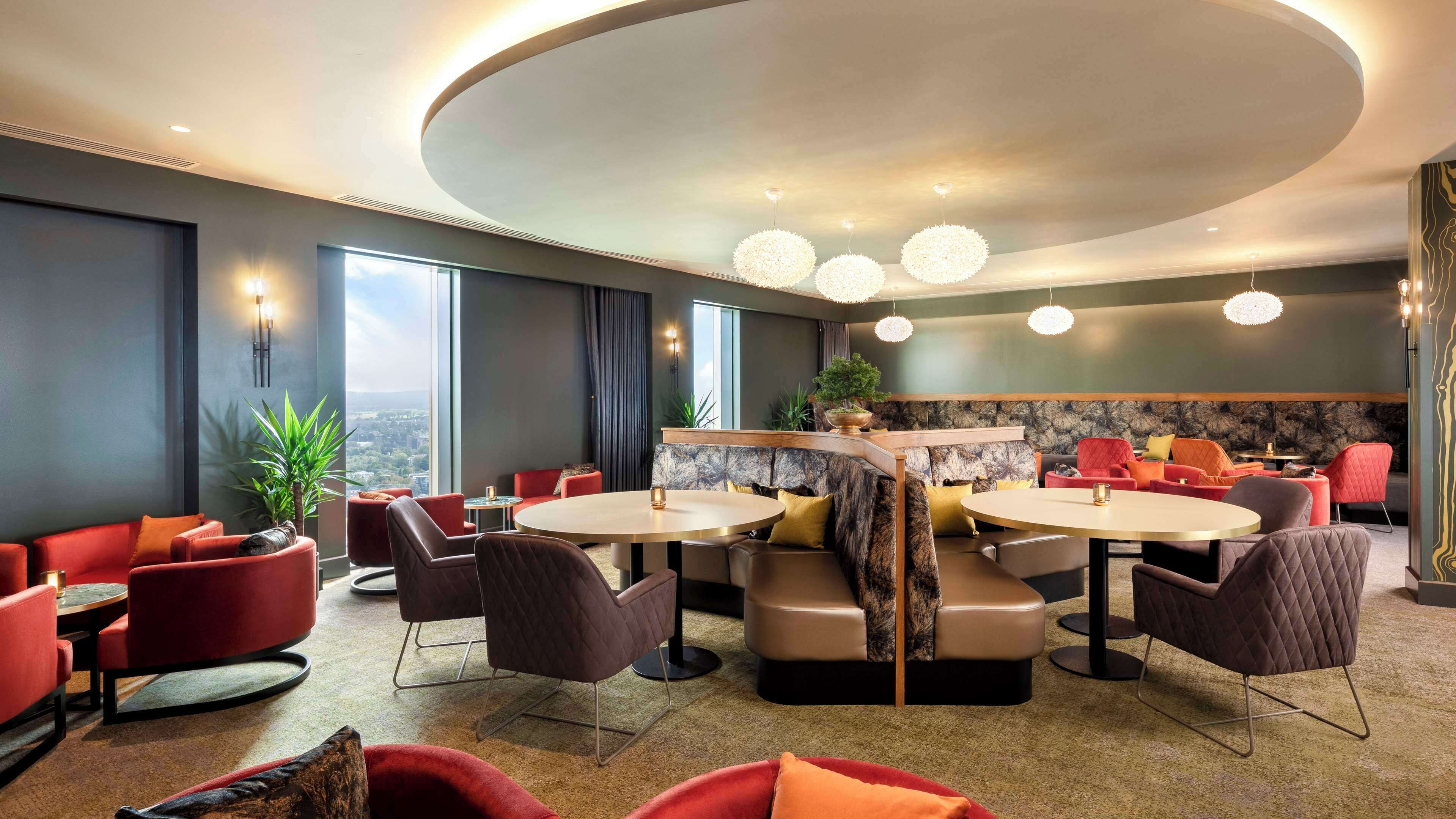 A modern lounge with round tables, red and brown seating, and a city view.
