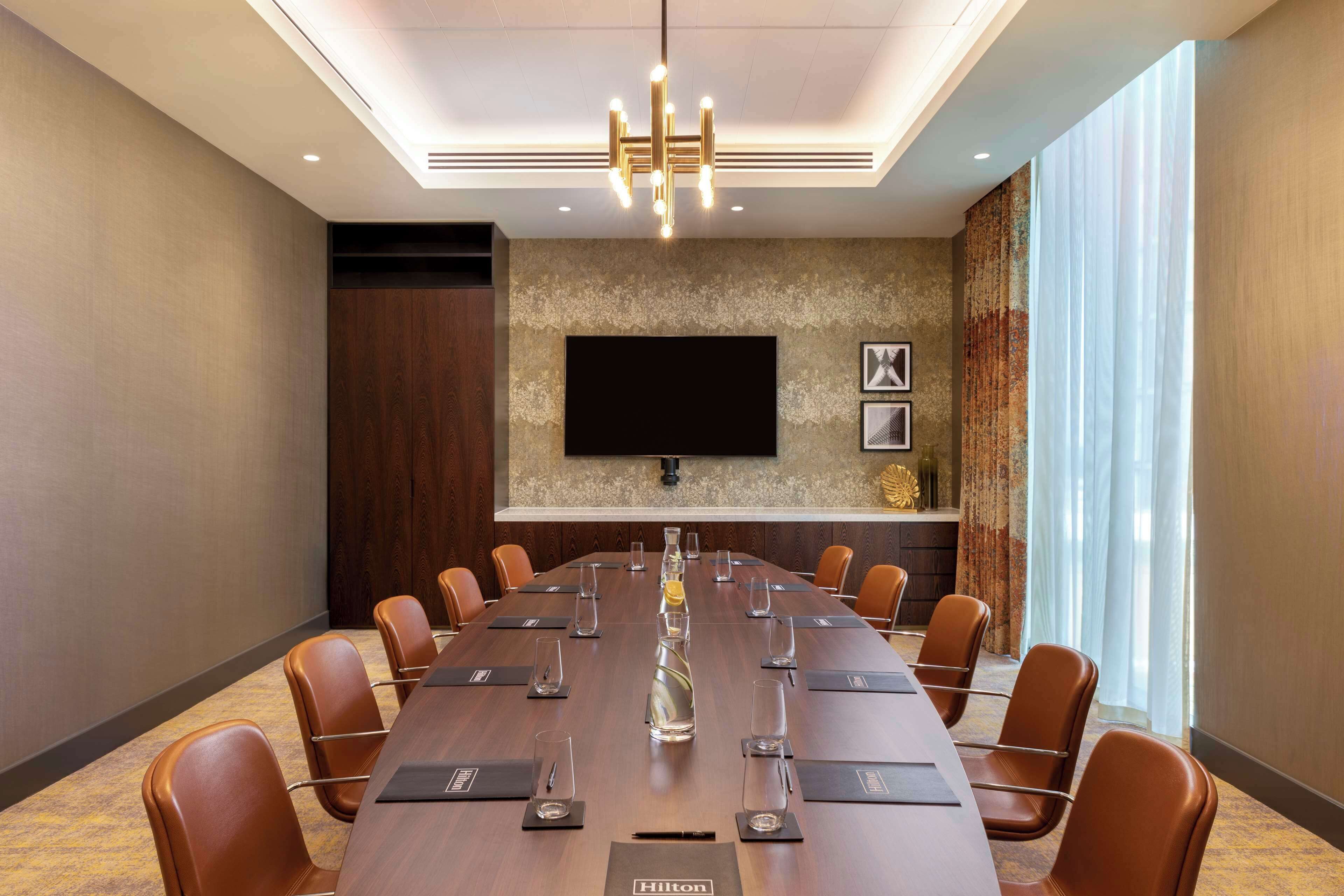 Ein Konferenzraum mit einem langen Tisch, braunen Ledersesseln, einem großen Bildschirm und einer modernen Chandelier.