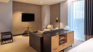 A modern hotel room with a sofa, a wooden side table, a flat-screen TV mounted on the wall, a small round table, and a large window with curtains.