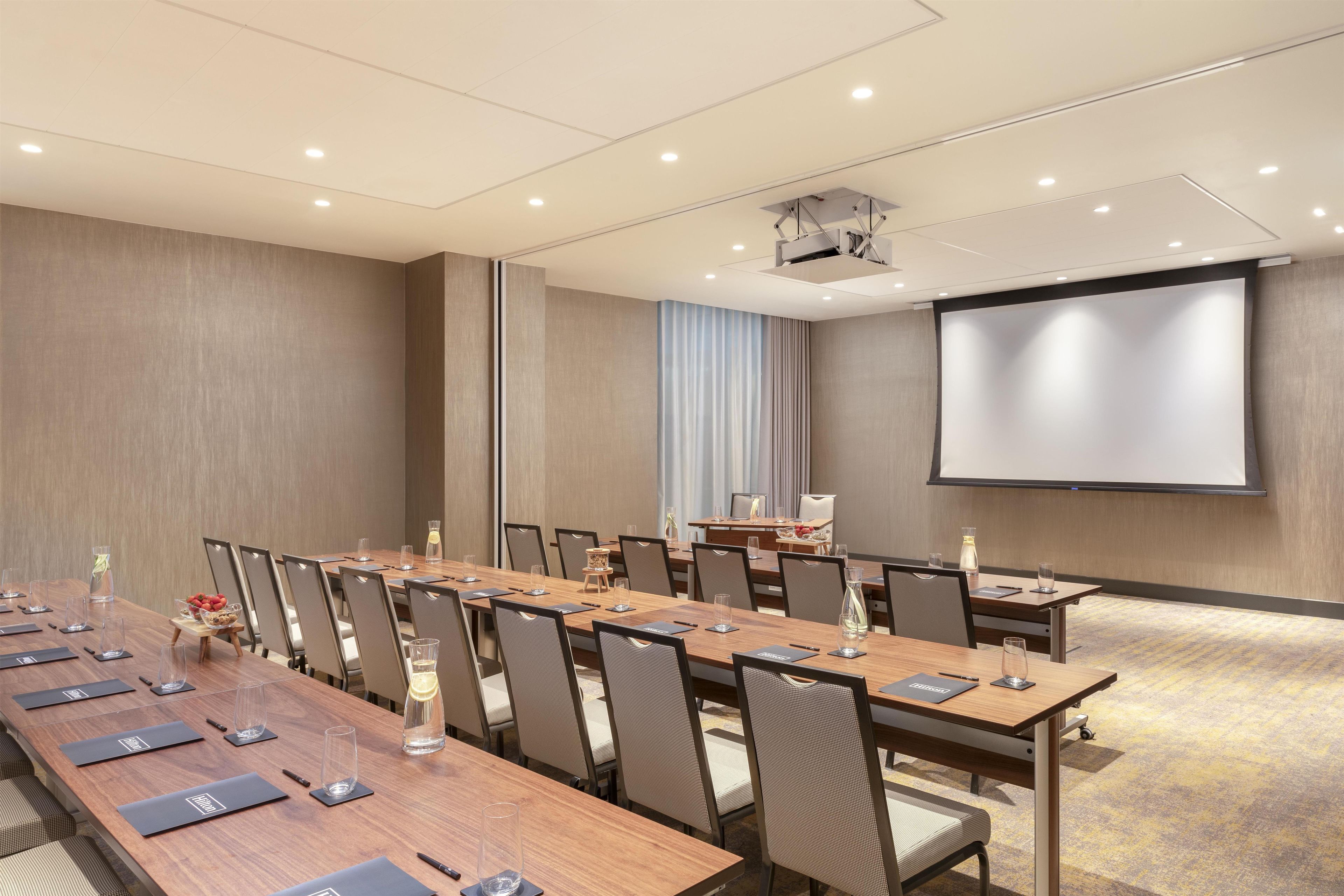 A conference room with a long table, chairs, a projector screen, and a screen for presentations.