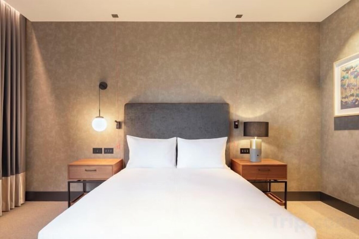 A hotel room with a large bed, two bedside tables, a nightstand, a lamp, and a wall-mounted light fixture.