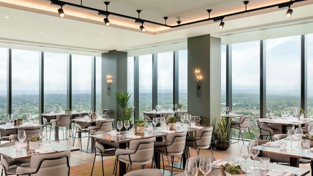 Un comedor moderno con mesas dispuestas para una comida, ventanas amplias que ofrecen vista a la ciudad y un techo alto con iluminación empotrada.
