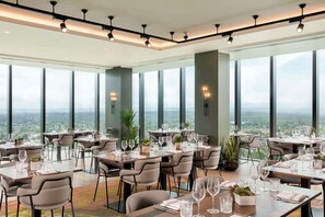 A modern dining area with tables set for a meal, large windows offering a city view, and a high ceiling with track lighting. - Hilton Woking (Woking)