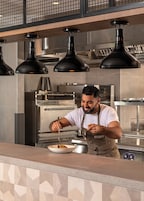 Un chef en una cocina moderna con iluminación industrial y electrodomésticos de acero inoxidable.