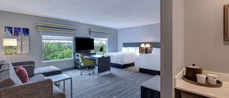 A hotel room with a sofa, two beds, a desk, a TV, and a view of greenery.