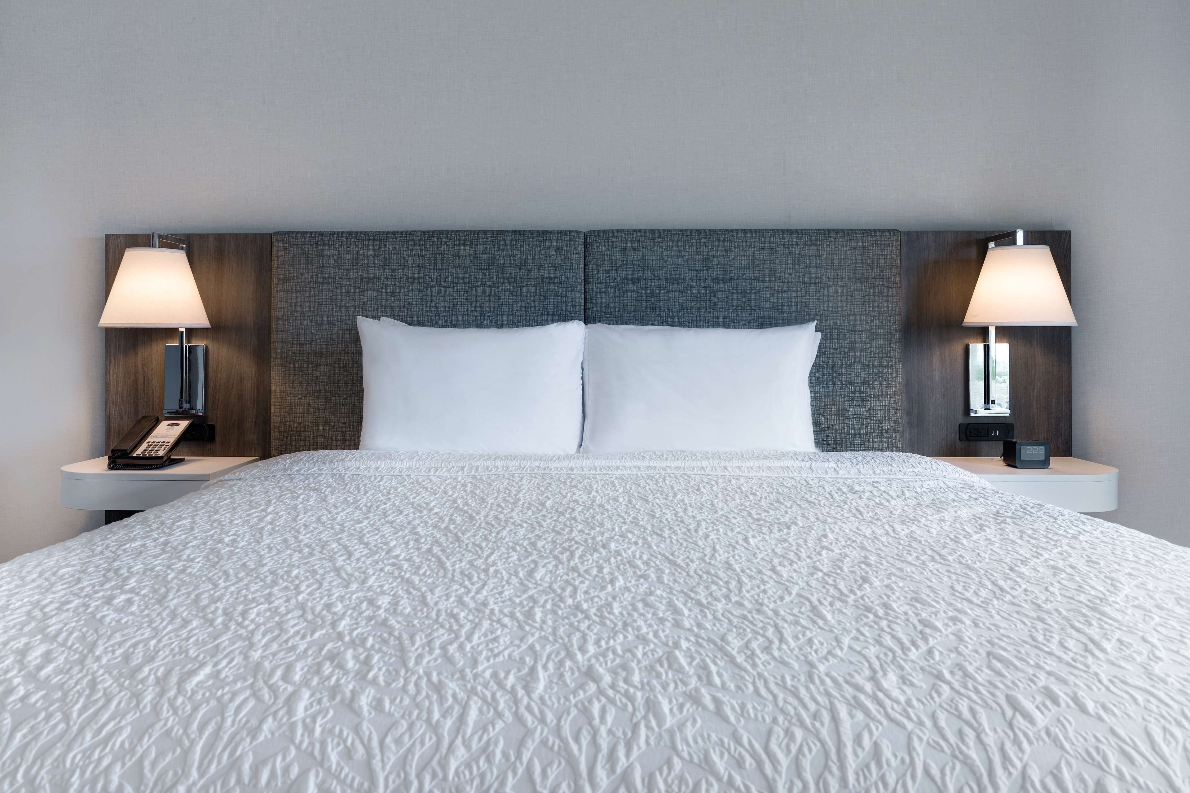 Una cama bien hecha con edredón blanco texturizado, dos mesitas de noche con lámparas y un cabecero moderno.