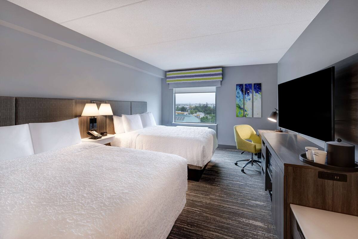 A hotel room with two beds, a desk, a TV, and a window with a city view.