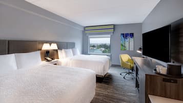 Ein Hotelzimmer mit zwei Betten, einem Schreibtisch, einem Fernseher und einem Fenster mit Blick auf die Stadt.