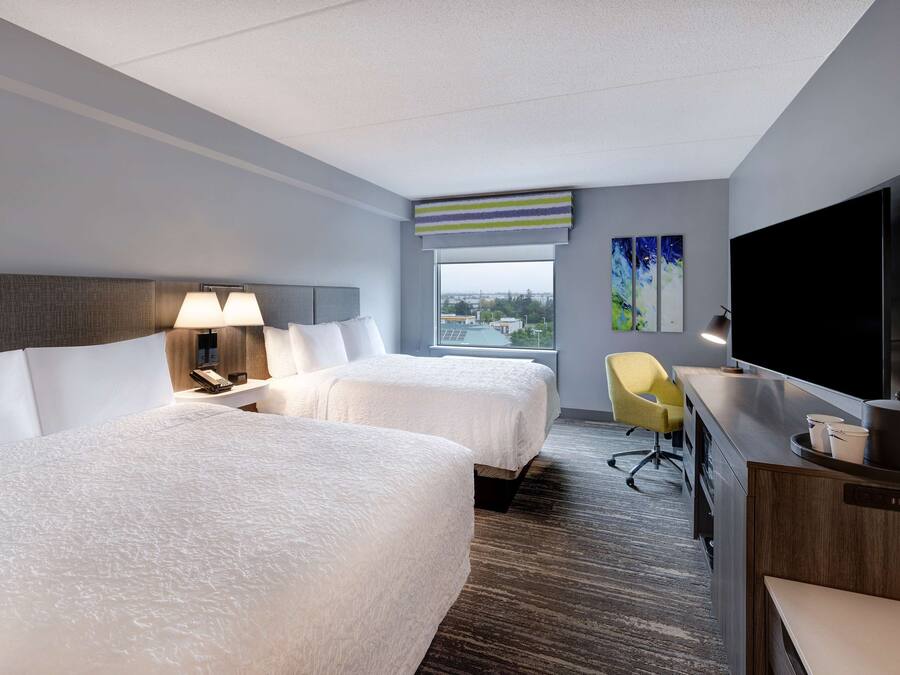 A hotel room with two beds, a desk, a TV, and a window with a city view.