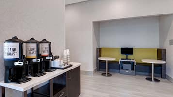 Un espace dédié au café, équipé de trois distributeurs de café étiquetés « Café torréfié foncé », « Café torréfié léger » et « Eau chaude ». On y trouve également deux petites tables et un téléviseur à l’arrière-plan.