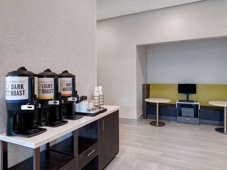 Un espace dédié au café, équipé de trois distributeurs de café étiquetés « Café torréfié foncé », « Café torréfié léger » et « Eau chaude ». On y trouve également deux petites tables et un téléviseur à l’arrière-plan.