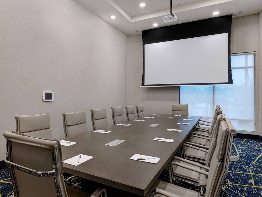Un salón de conferencias con una larga mesa, varias sillas, una pantalla de proyector y un ventanal grande.