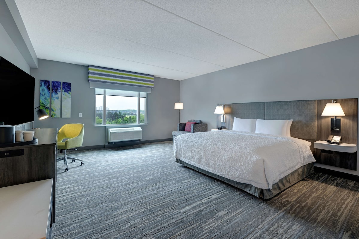 A hotel room with a large bed, a desk, a chair, a window with a view, and a lamp.