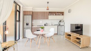 Eine moderne Küche mit weißen Schränken, ein Essbereich mit einem runden Tisch und weißen Stühlen, ein Fernseher auf einem Holzboden und ein Topfpflanze.