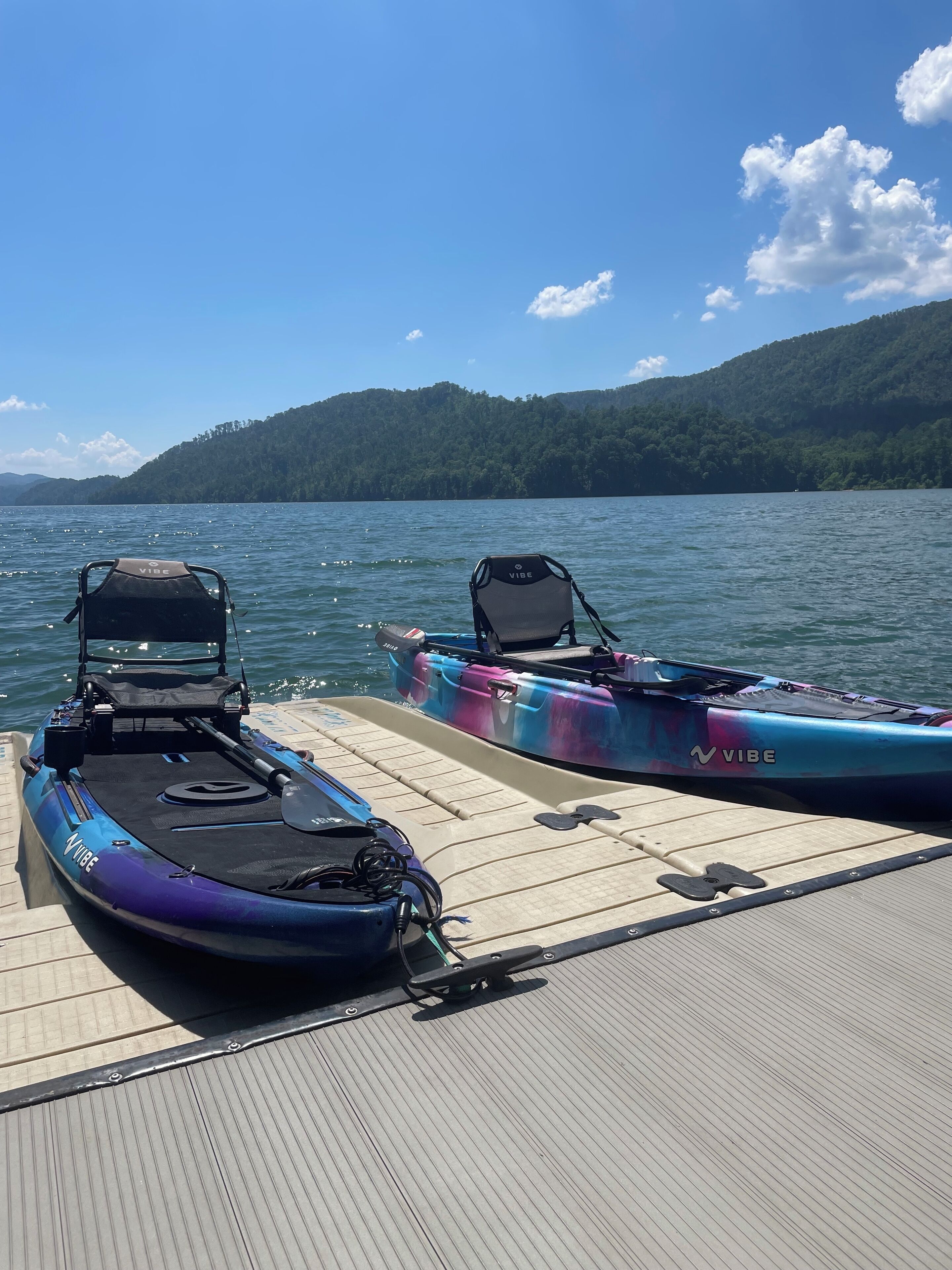 Two kayaks and a paddleboard on a wooden dock by a lake with a forested hill in the background.