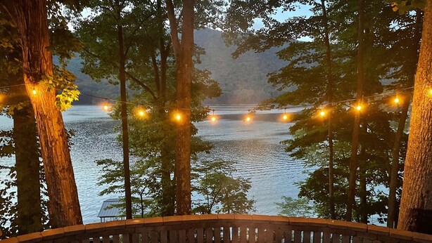 Un foyer en pierre, entouré d’un banc en pierre incurvé, surplombe un lac illuminé de guirlandes lumineuses et bordé de grands arbres.