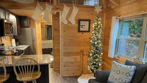 L’intérieur chaleureux d’un chalet comprend un coin cuisine avec des armoires en bois, un comptoir en granit et un réfrigérateur. On peut apercevoir un sapin de Noël décoré de guirlandes lumineuses et d’ornements, et un tableau encadré accroché au mur porte l’inscription : « Les meilleurs souvenirs se créent au bord du lac Watonga. »