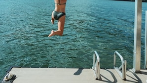 Una mujer salta a un lago desde un muelle, con una montaña boscosa al fondo.