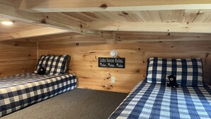 Une chambre chaleureuse dans un chalet, avec deux lits, chacun recouvert d’une couverture à carreaux et agrémenté d’un petit ours en peluche. Une pancarte sur le mur indique : « Règles de la maison au bord du lac : détendez-vous… détendez-vous… détendez-vous. »