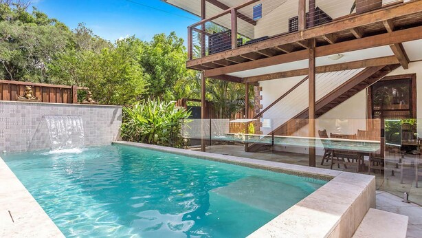 Une piscine dotée d’une terrasse en bois et d’une barrière en verre, entourée d’une végétation luxuriante et d’un mur de pierre.