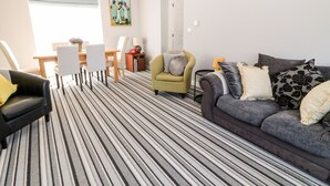 A modern living room with a striped carpet, a sofa, a black armchair, and a dining area with a table and chairs. - Seagull Cottage (Bude)