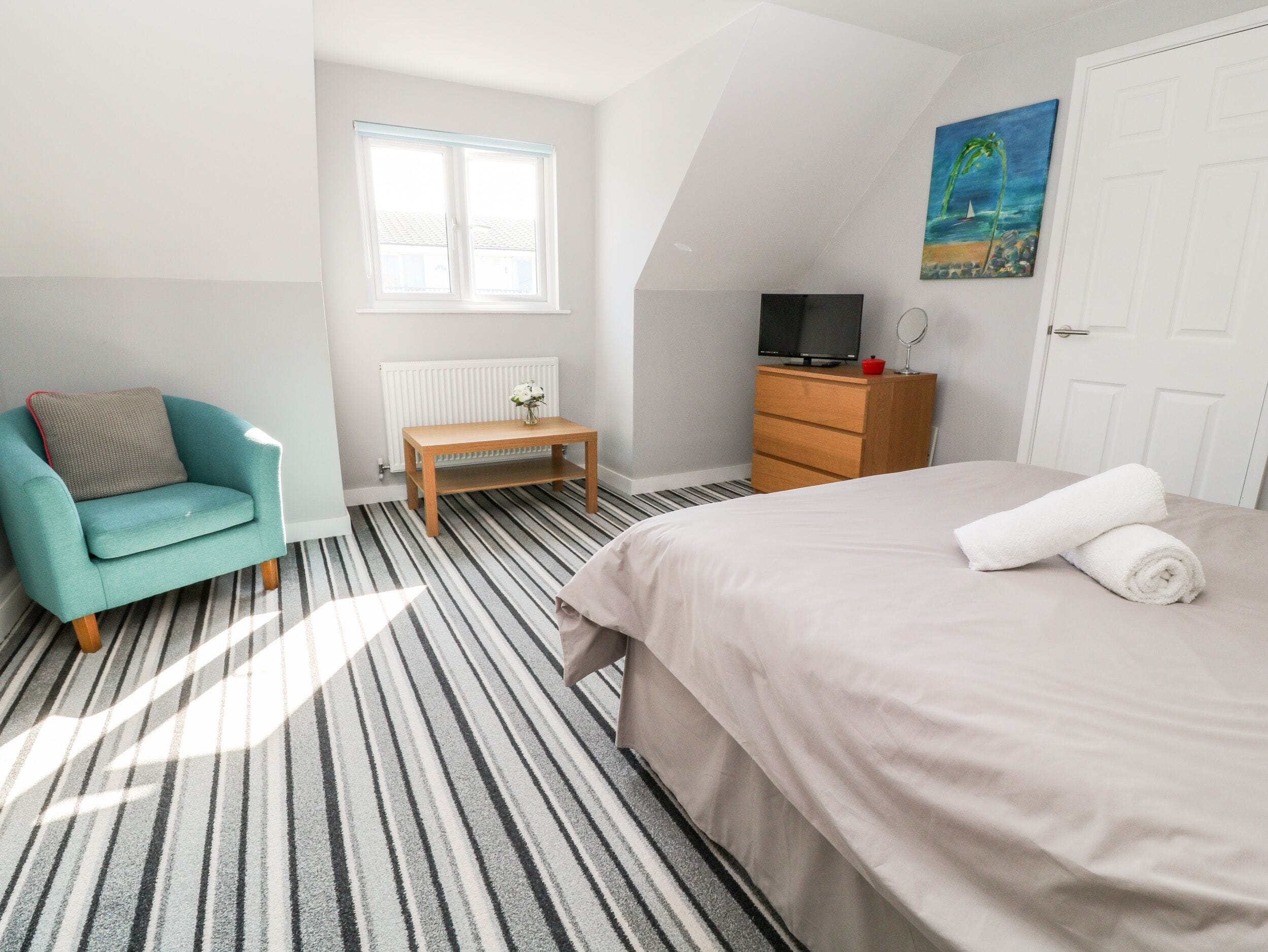 A bedroom with a striped carpet, a bed with a rolled towel, a teal armchair, a wooden dresser, a television, and a window with blinds.