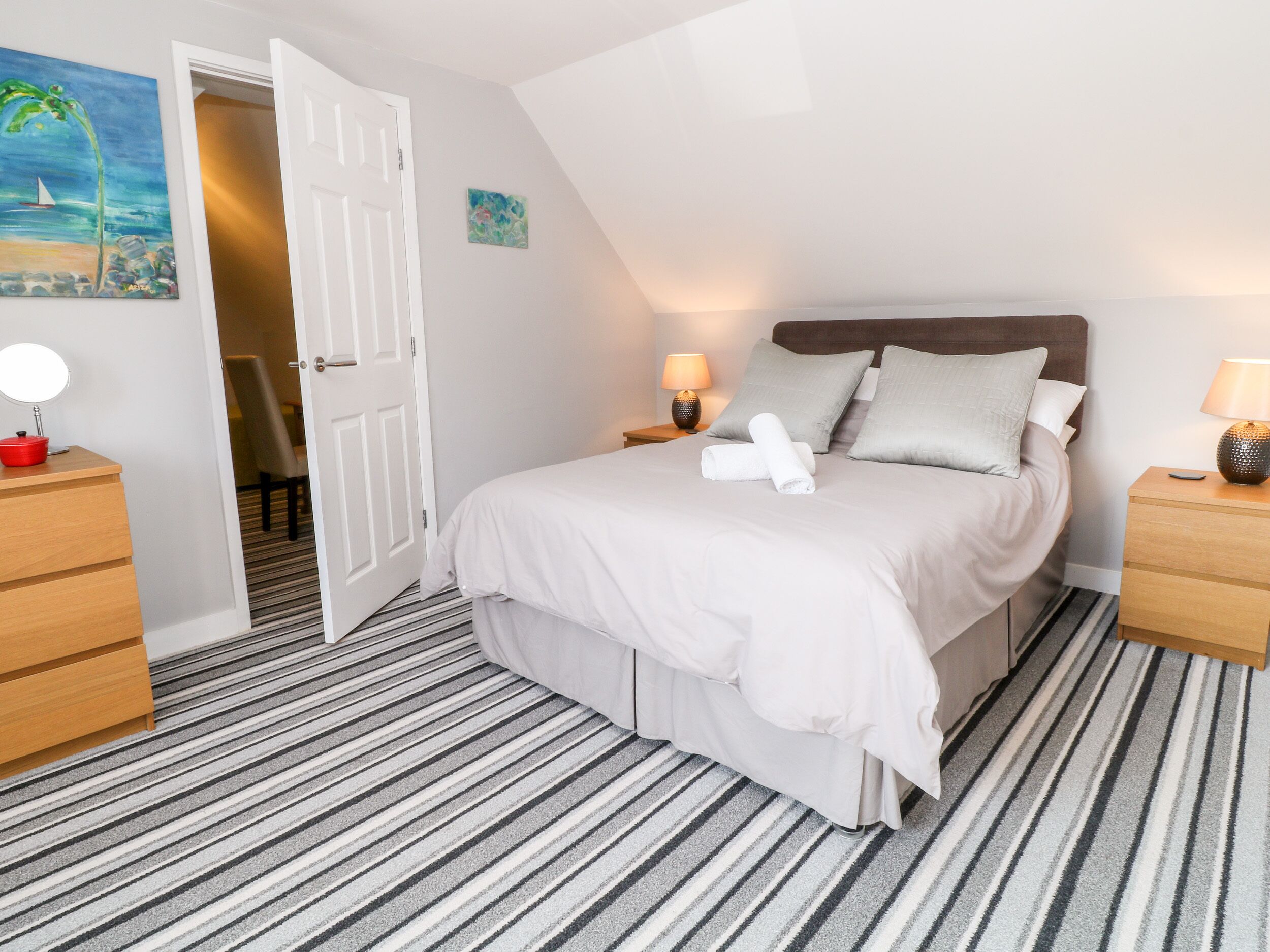 Un dormitorio ordenado con una cama, dos mesitas de noche, una puerta que da a otra habitación y una alfombra a rayas.