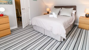 A neatly arranged bedroom with a bed, two bedside tables, a nightstand, a door leading to another room, and a striped carpet. - Seagull Cottage (Bude)