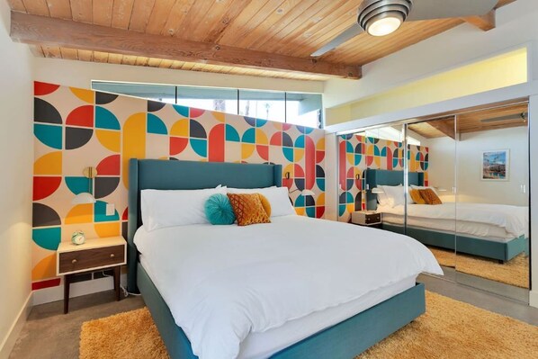A modern bedroom with a large bed, a nightstand, a mirror, and a colorful abstract wall design.