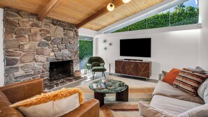 A modern living room with a stone fireplace, comfortable seating, a flat-screen TV, and a large window overlooking greenery.