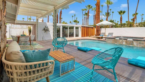 A pool area with lounge chairs, a wooden table, and a covered seating area with a view of palm trees.