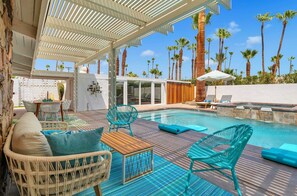 A pool area with lounge chairs, a wooden table, and a covered seating area with a view of palm trees.