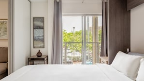 Ein Hotelzimmer mit einem großen Bett, einem kleinen Tisch, einer Vase und einem Gemälde. Es gibt einen Balkon mit Blick auf Bäume und Gebäude.