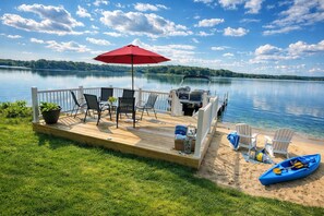 A wooden deck with outdoor furniture, a red umbrella, and a kayak by the lake. - PONTOON BOAT NOW INCLUDED! KAYAKS! Waterfront Cottage on Mona Lake with HOT TUB! (Norton Shores)