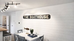 Un coin repas avec une table blanche, des chaises grises, une décoration représentant un poisson et une enseigne sur le mur indiquant « THE BOATHOUSE ».