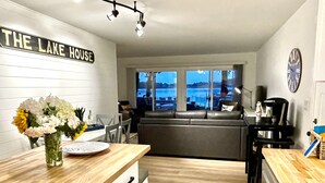 Une cuisine moderne avec un plan de travail en bois, un coin repas avec une table et des chaises, une grande fenêtre donnant sur le lac et une enseigne indiquant « THE LAKE HOUSE ».