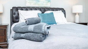 Ein ordentlich bezogenes Bett mit einer grauen und weißen Tagesdecke, einem blauen Kopfkissen und einem Stapel gefalteter grauer Handtücher.