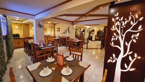 Uma sala de jantar com mesas e cadeiras de madeira, uma cozinha ao fundo e um desenho decorativo de uma árvore na parede.